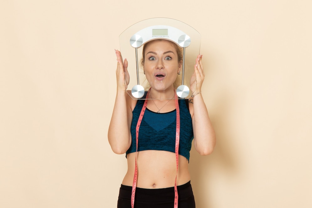 A young woman in a sporty outfit holding a digital scale above her head with a surprised expression. She is wearing a blue crop top, black leggings, and a pink measuring tape around her neck.