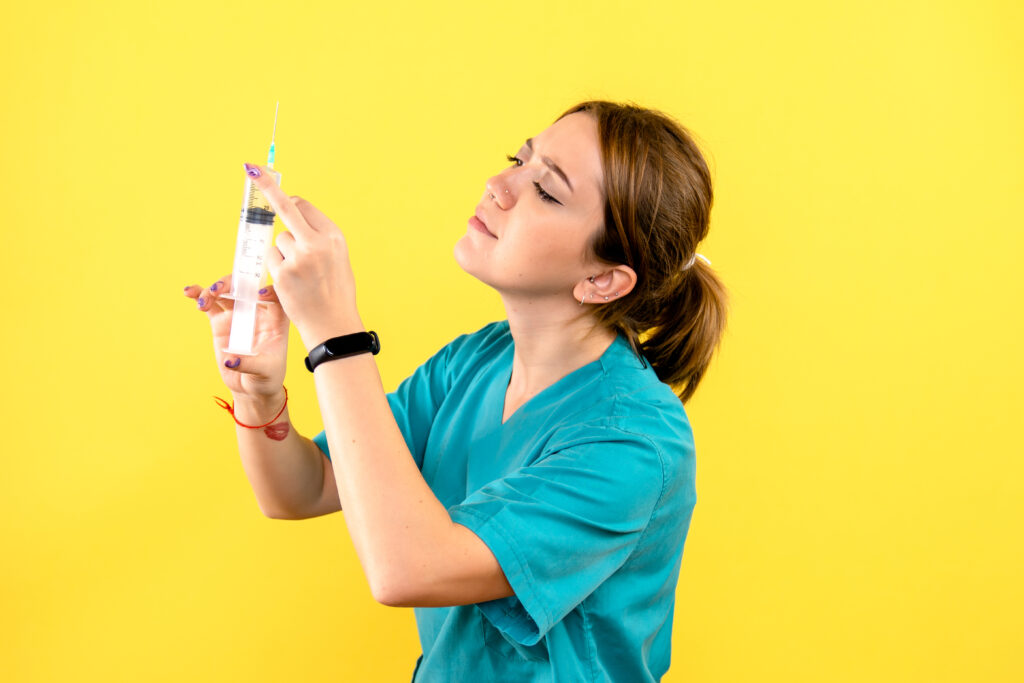 A female healthcare worker in teal scrubs holding a syringe and preparing an injection against a bright yellow background.